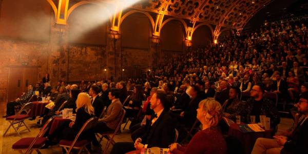 DinnersAndGalas_masthead_image The grand hall with orange-tinted lighting. People sit at cabaret-style tables at the front, with raked seating behind all the way to the back of the hall