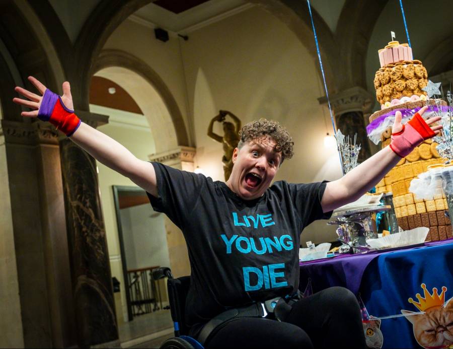 live young die biscuity Jess Thom, a woman with short brown curly hair, holds her arms wide in front of a table decorated with cats-in-party-hats bunting and a giant tiered biscuit installation. Photo by Samuel Dore.