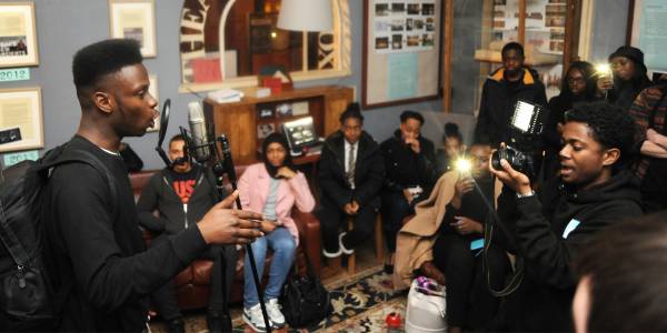 AgencyforYoungPeople_Masthead_Image A group of around 15 young people gather in a room, sitting on sofas and chairs. Some are taking photos with their phones. In the foreground one person talks into a microphone with a pop guard, gesturing with his hand, and another points an SLR camera at him
