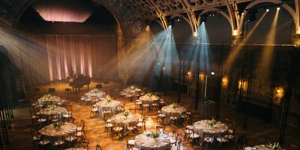 BAC_-_Wedding_--_Commercial_Edit-43 An image of the Grand Hall set up for a wedding. There are lines of circular tables dressed with white table-cloths and flower bouquets. There is a grand piano at the far end of the room and spotlights shining down from the ceiling.