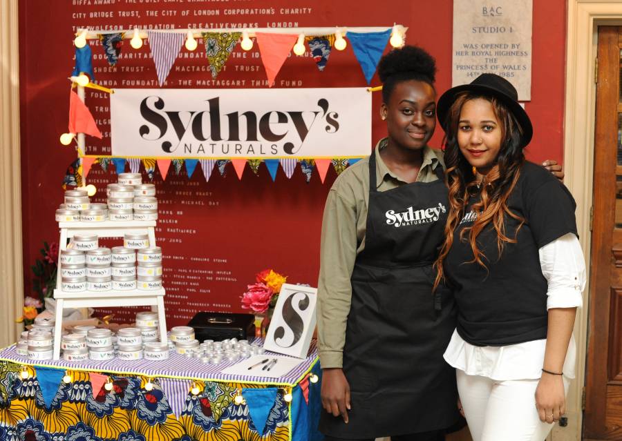 The Agency Against a red wall, a stall with colourful bunting is set up for Sydney's Naturals, with lots of branded tins, a colourful tablecloth, and flowers. Sydney, wearing a black branded apron and green shirt, and another agent, wearing a black fedora, branded black t shirt, and white trousers, stand to the right of the stall