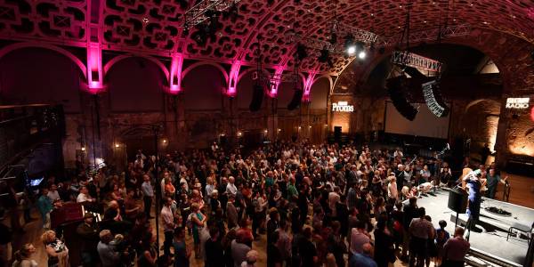Proms at Battersea Arts Centre_Hires_16.9 A large standing crowd in the Grand Hall, with the ceiling lit up pink. A stage in the middle of the right hand side has a musician and a harp. Projected onto the far wall are logos for BBC Radio and BBC Proms