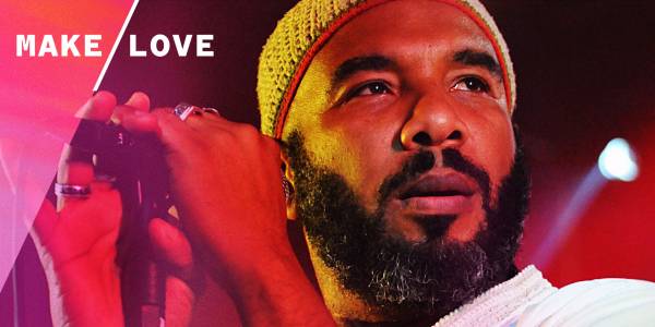 Anthony Joseph A close up image of a man with a dark beard under red stage lights holding a microphone wearing a beaded hat and white shirt.