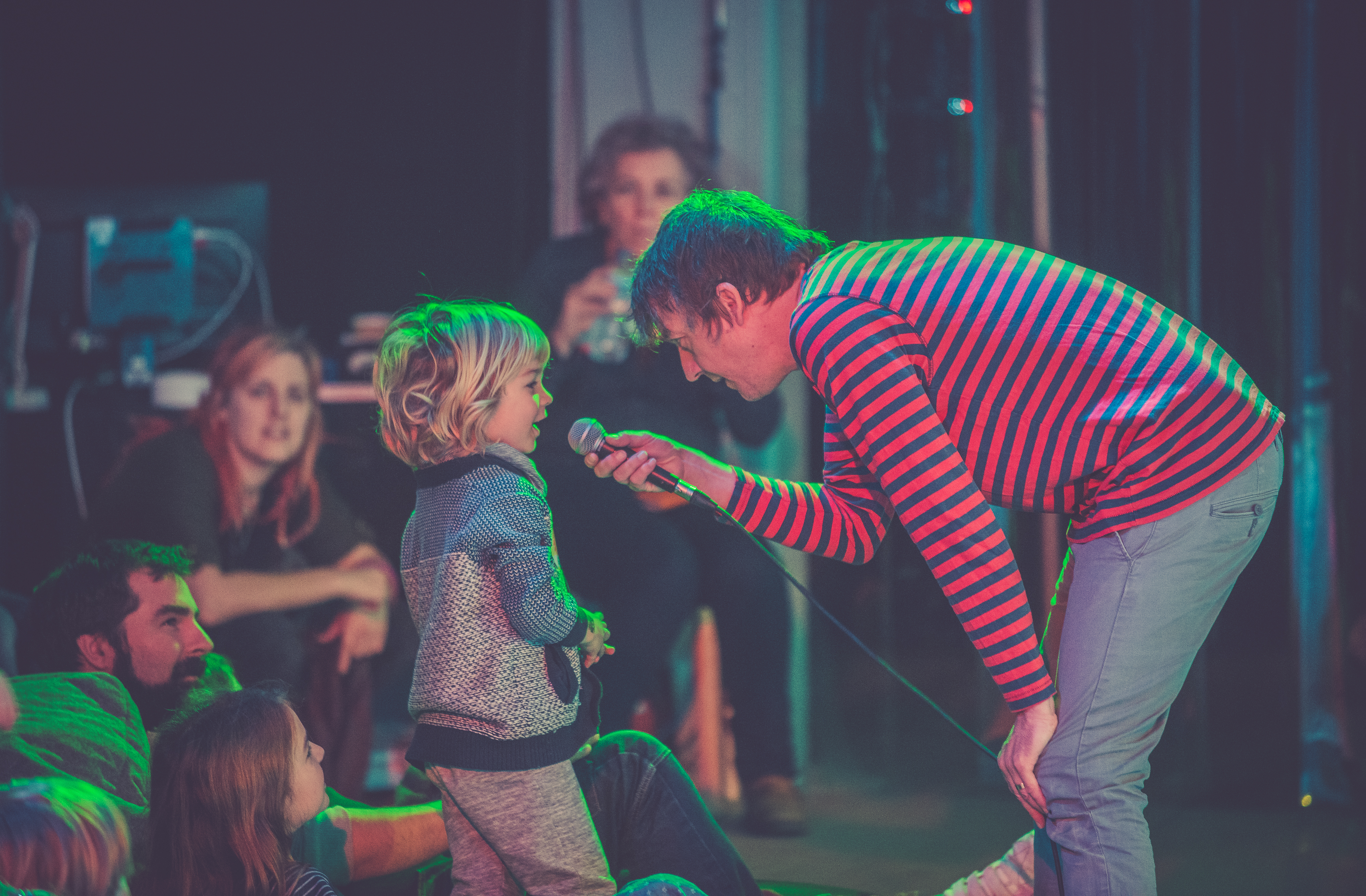 Moving Roots An image of an artist wearing a red and black striped long tshirt holds a microphone to a young child with blonde hair under green and pink flourescent lights.